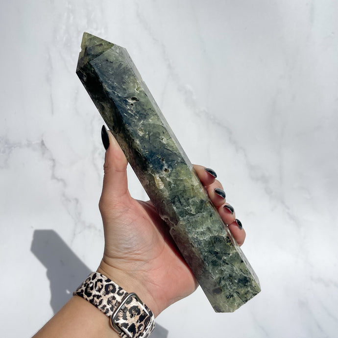 Hand holding a green Prehnite with Epidote crystal tower against a white marble background