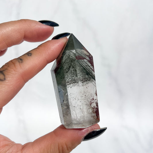 Person holding a quartz crystal with green phantoms and small red garden inclusions on a white background