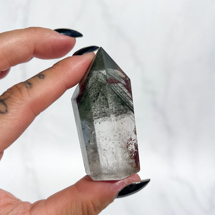 Person holding a quartz crystal with green phantoms and small red garden inclusions on a white background