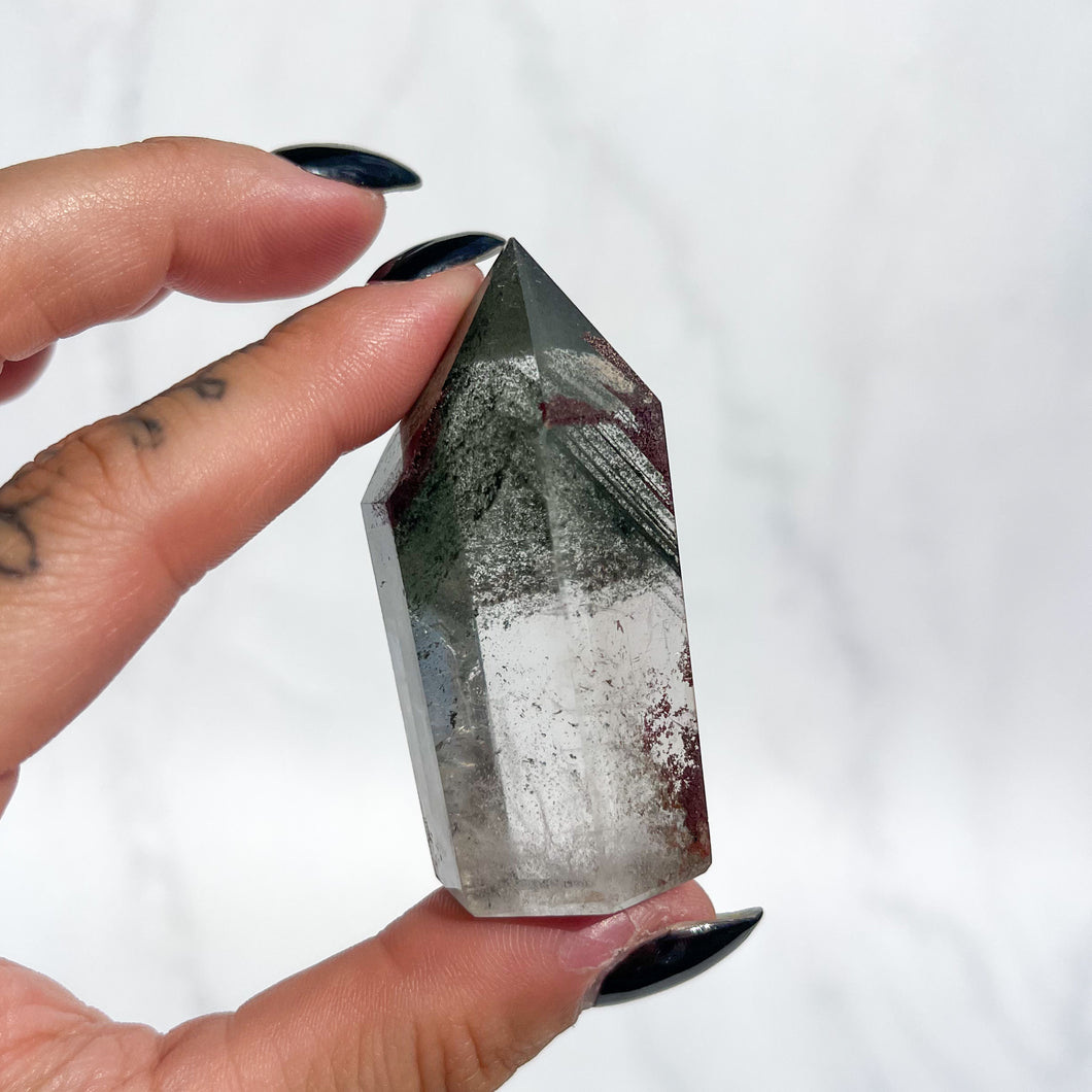 Person holding a quartz crystal with green phantoms and small red garden inclusions on a white background